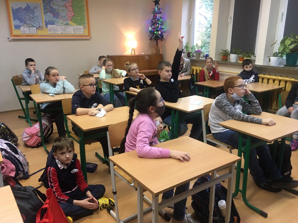 Sala lekcyjna. Uczniowie siedzą w ławkach. Jeden z uczniów podnosi do góry rękę.