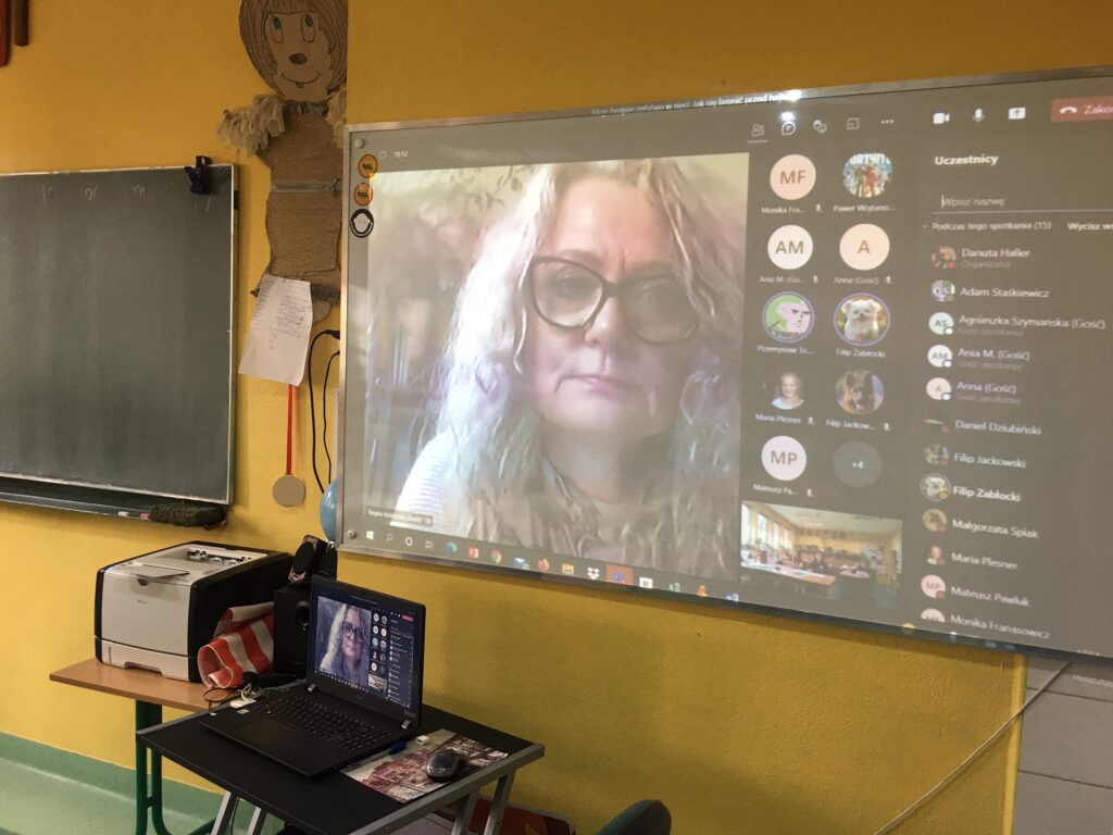 Udostępniony obraz na tablicy interaktywnej. Po lewej stronie zdjęcie prowadzącej . Na dole po prawej stronie widok uczniów w klasie, uczestniczących stacjonarnie na lekcji w klasie. Po prawej stronie uczestnicy tej lekcji online

