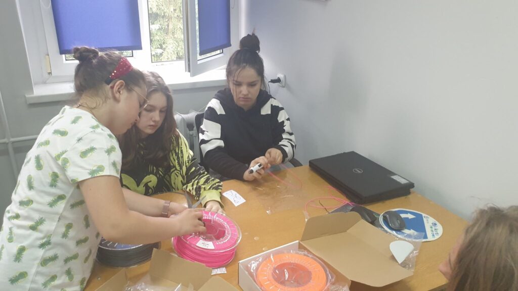 Na zdjęciu widoczni uczniowie, którzy przygotowują się do pracy z długopisem 3D. Wybierają kolor filamentu i rozpoczynają pracę. 