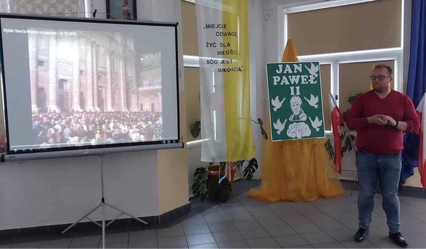 Hol szkoły. Na środku mężczyzna prowadzący spotkanie. W tle ekran z wyświetlanym materiałem merytorycznym.