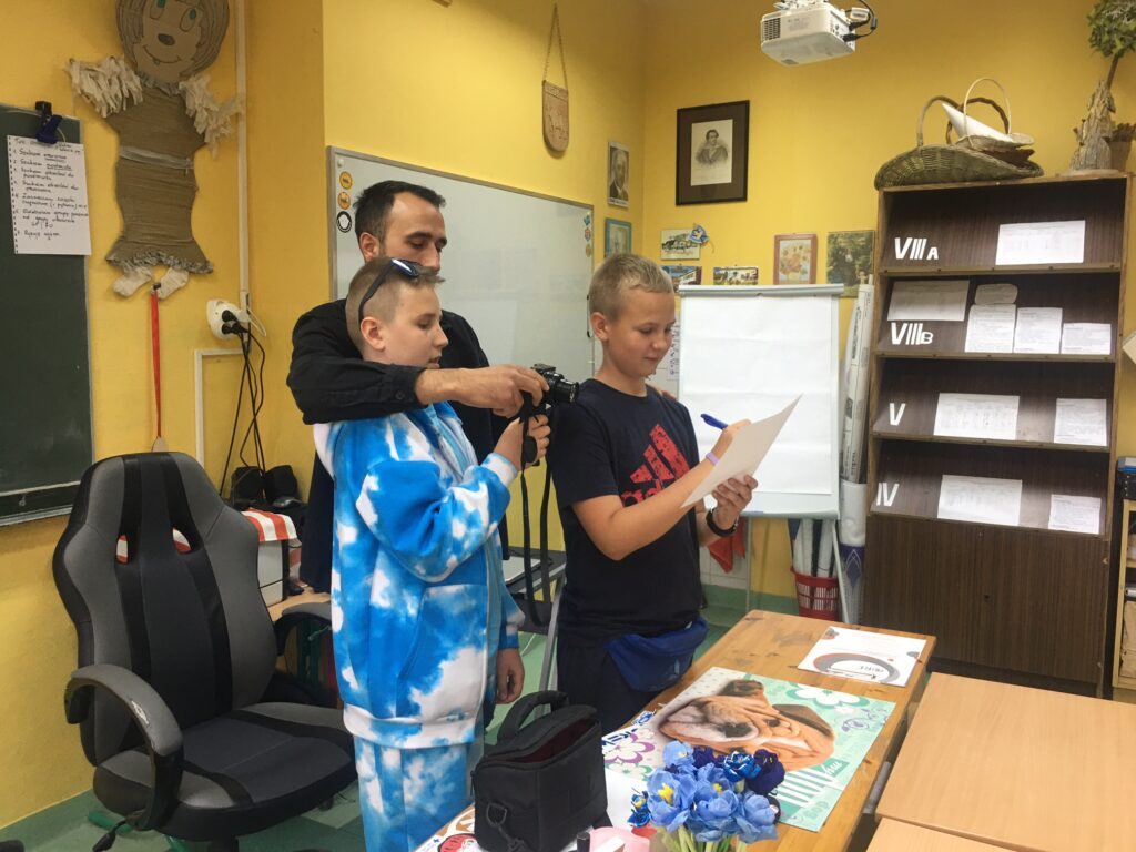 Nauczyciel w czarnym ubraniu stoi przed biurkiem za dwoma chłopcami, którym coś tłumaczy. Jeden chłopak ubrany na niebiesko ma w ręku aparat fotograficzny a drugi w czarnej koszulce pisze coś na białej kartce.

