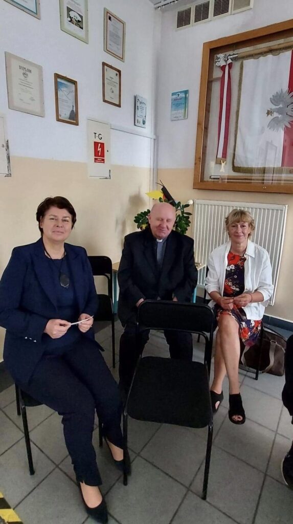 Na tle białej ściany siedzą dwie kobiety i w środku ksiądz.