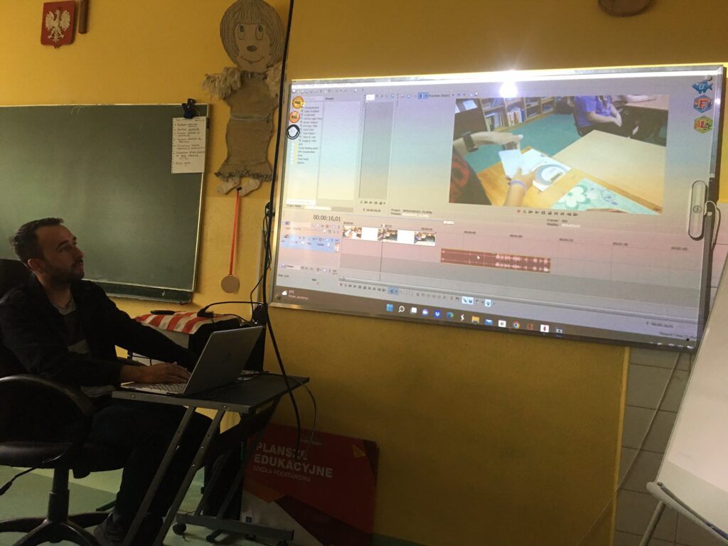 Sala lekcyjna. Na wprost ekran z pokazanym fragmentem nagranego filmiku. Po lewej stronie siedzi przed laptopem ubrany na czarno nauczyciel. 

