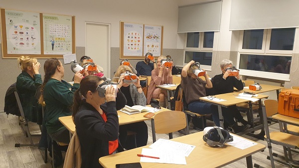 Na zdjęciu widoczna grupa nauczycieli, siedzą w ławkach. Mają założone okulary VR i patrzą przez nie. 
