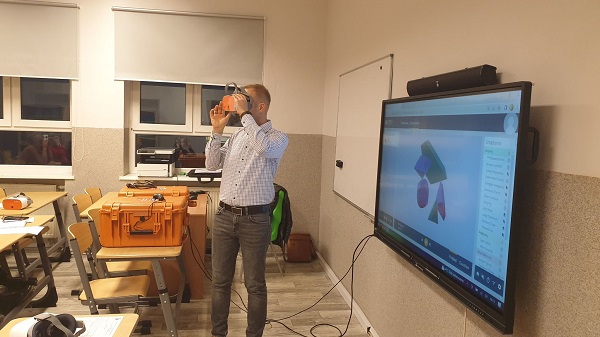 Na zdjęciu widoczny mężczyzna, który stoi, ma założone okulary VR. Z prawej strony wisi monitor interaktywny, z lewej ławki. 