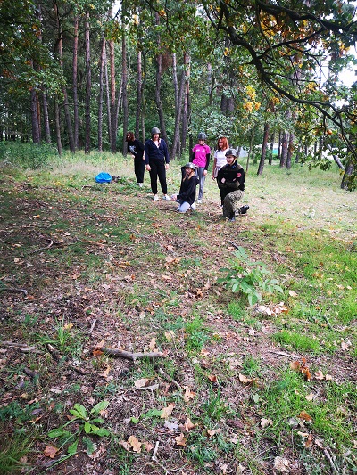 grupa 5 dziewczynek na tle lasu przygotowuje się do konkurencji rzutu granatem, jedna dziewczynka klęczy na jedno kolano i wykonuje zamach ręką prawą, pozostałe stoją kilka metrów za nią w hełmach na głowie