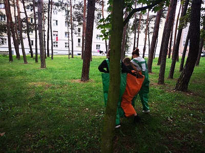trzy dziewczynki ubrane w gumowe zielono - pomarańczowe spodnie, które są ze sobą połączone nogawkami próbują pokonać kilkunastometrowy dystans między drzewami