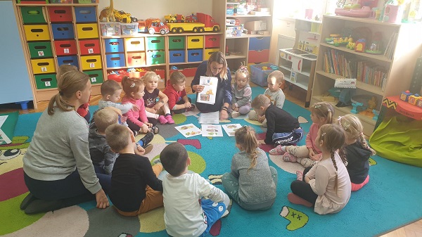 Na zdjęciu widoczne dzieci i prowadząca zajęcia nauczycielka. Wszyscy siedzą na dywanie. Nauczycielka pokazuje dzieciom obrazki ze zdrową żywnością i czyta wierszyk. 
