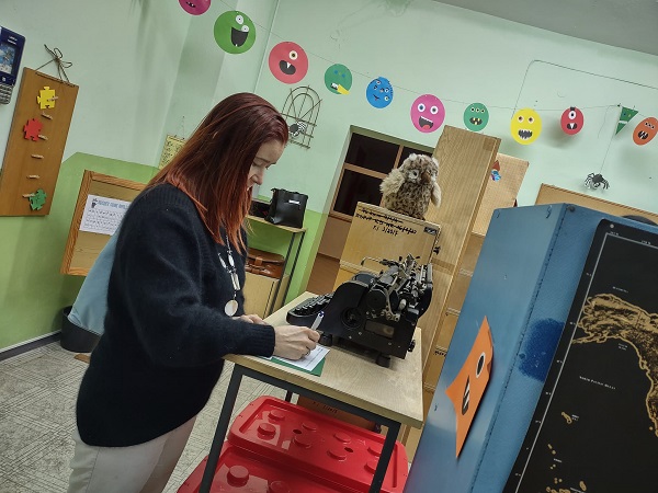 Na zdjęciu widoczna nauczycielka, która rozwiązuje zadanie. Jest w pokoju zagadek, za nią stoi maszyna do pisania. 
