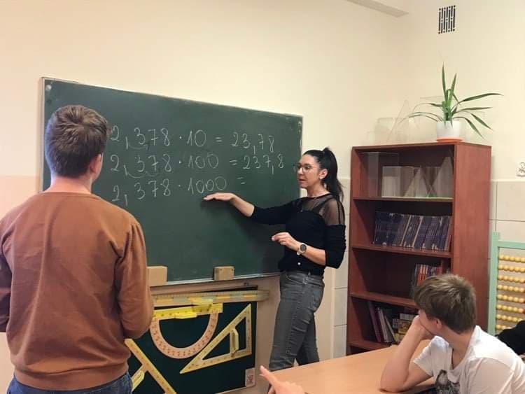 Na zdjęciu widoczny nauczyciel, który stoi przy tablicy i tłumaczy uczniom mnożenie ułamków. 