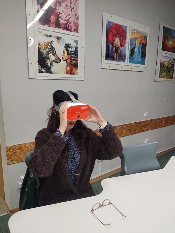 Na zdjęciu nauczycielka siedząca na krześle przy stole, która ma założone okulary VR.

