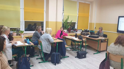 Sala lekcyjna, przy stolikach siedzą nauczyciele. Na ścianie ekran z prezentacją. Po lewej stronie przy biurku siedzi osoba prowadząca. Ubrana jest w zieloną bluzką. Po lewej stronie okno przysłonięte żółtymi roletami. 