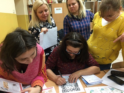 Sala lekcyjna, przy stoliku siedzą dwie kobiety i stoją trzy kobiety – nauczycielki, które maja przed sobą kolorowe kartki z zadaniami do rozwiązania. Wskazują coś palcami i jedna z nich zapisuje dane rozwiązanie  na kartce.
