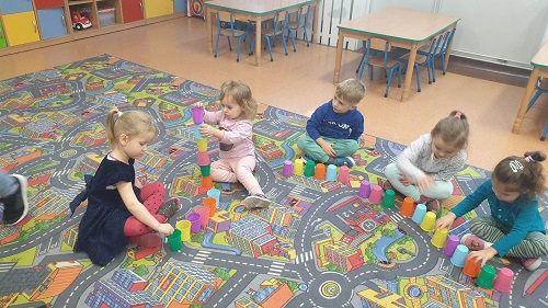 Na zdjęciu widoczne dzieci. Cztery dziewczynki i jeden chłopiec, siedzą na dywanie. Przed każdym stoi 6 kubeczków w różnych kolorach. Próbują budować wieżę. 