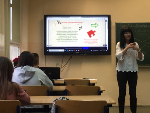 Sala lekcyjna, z przodu ekran z kolorową prezentacją, po jego prawej stronie zielona tablica do pisania. Przed monitorem stoi kobieta w białej bluzce i czarnych spodniach. W ławce, przed nią siedzą uczniowie. 

