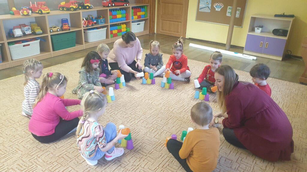 Dzieci siedzą na dywanie w kółku. Pomiędzy nimi siedzą również dwie Panie. Przed każdym dzieckiem stoi zestaw 6 kolorowych kubeczków. Dzieci budują wieżę, na wzór wieży zbudowanej przez nauczycielkę. 