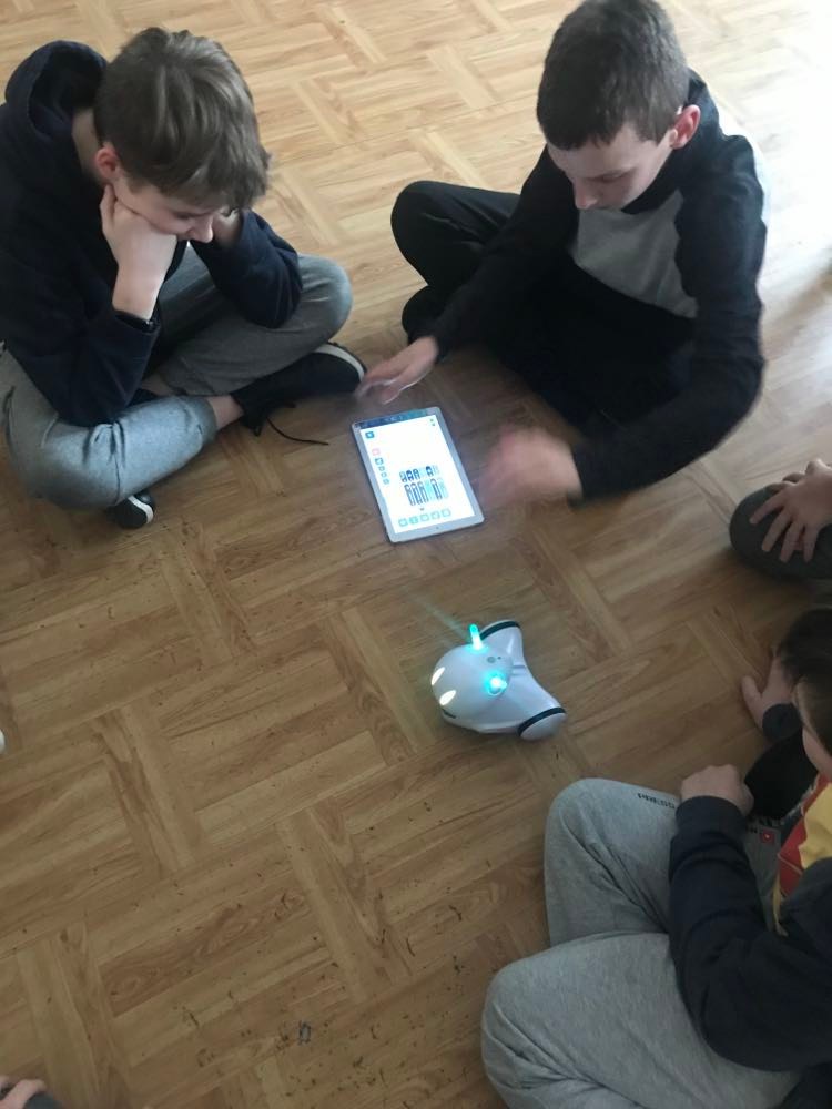 Na zdjęciu widocznych trzech uczniów, siedzą na podłodze. Przed nimi leży tablet i robot Photon.