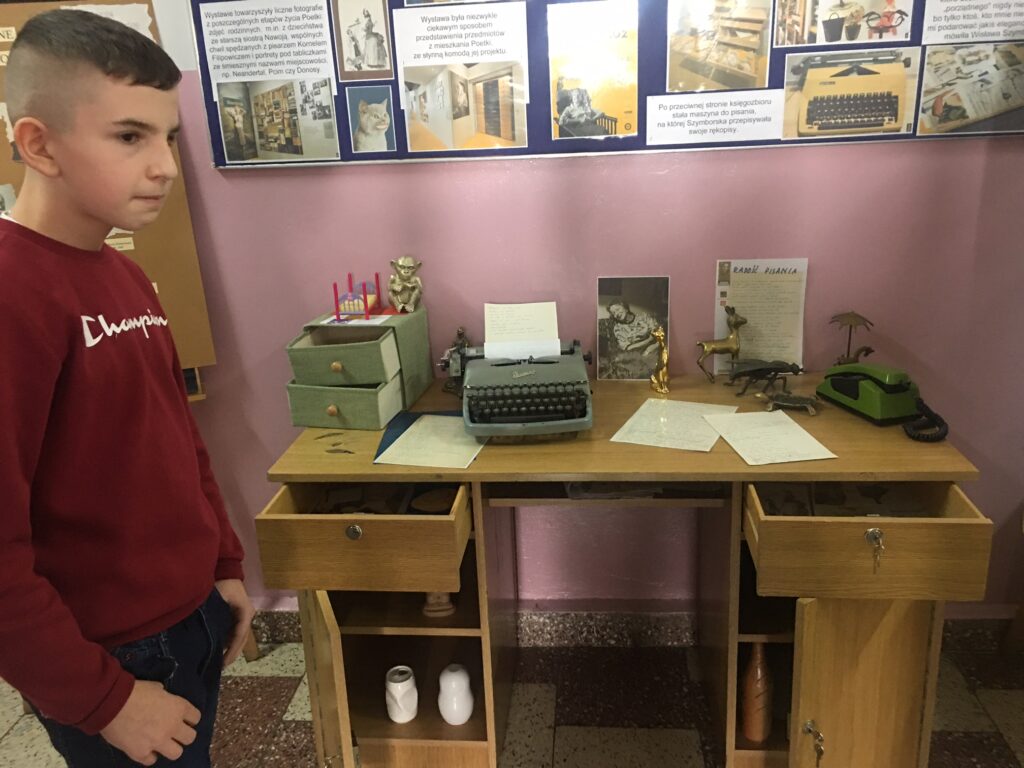Biurko, na którym jest maszyna do pisania, popielniczka, koty. Z lewej strony chłopiec w czerwonym sweterku