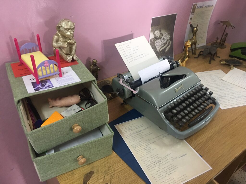 Biurko, na którym jest maszyna do pisania, zdjęcie trzyszufladowy pojemnik zielony z przedmiotami. Z boku figurka kota.