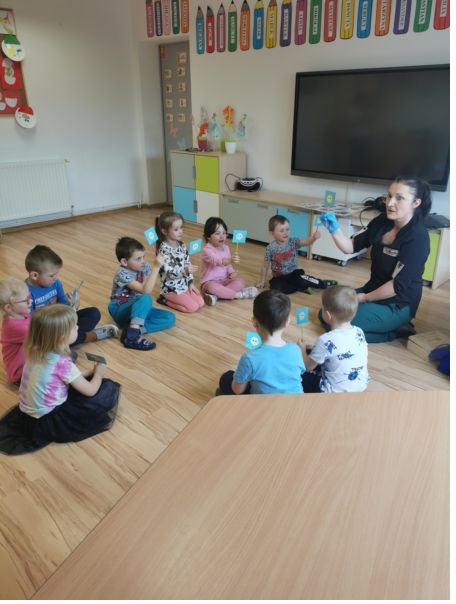 Dzieci siedzą w kole na podłodze. Nauczycielka w czarnej bluzce siedzi wśród dzieci .
Dzieci trzymają w rękach niebieskie kartoniki na patyczkach, uniesione do góry.
W tle na ścianie  wisi ekran , nad nim dekoracja kolorowe kredki.
