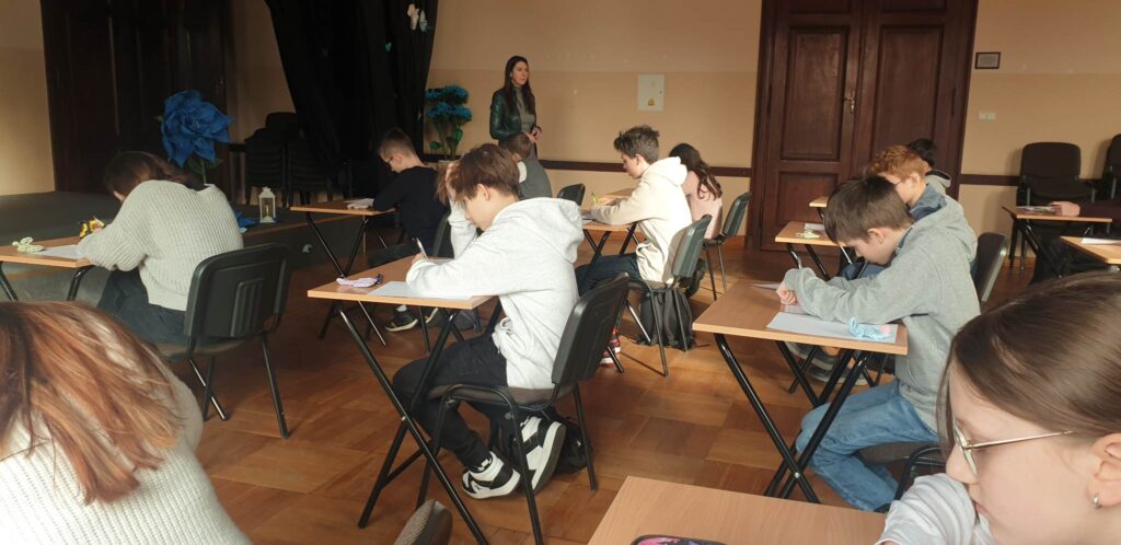 Na zdjęciu widoczni uczniowie, siedzą w ławkach, przed nimi leżą kartki z zadaniami, rozwiązują. 