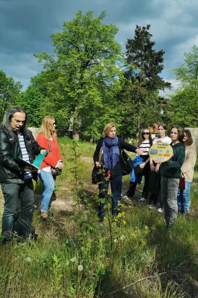 Fotografia przedstawia grupę osób stojących naprzeciwko siebie na zielonej polanie. Mężczyzna po lewej przegląda katalog z fotografiami, dwie kolejne osoby stoją w centralnej części zdjęcia obok grupy czterech uczniów, z których jeden trzyma w rękach żółtą, owalną tablicę z napisem, że w tym miejscu stał dom rodziny Steinhardt.
