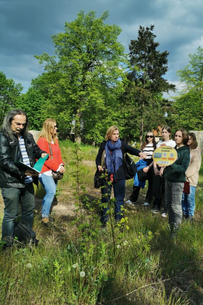 Fotografia przedstawia grupę osób stojących naprzeciwko siebie na zielonej polanie. Mężczyzna po lewej przegląda katalog z fotografiami, dwie kolejne osoby stoją w centralnej części zdjęcia obok  grupy czterech uczniów, z których jeden trzyma w rękach żółtą, owalną tablicę z napisem, że w tym miejscu stał dom rodziny Steinhardt. 