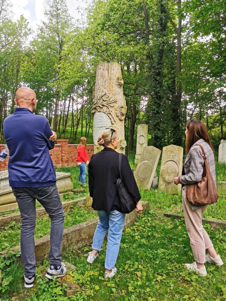 Na pierwszym planie fotografii znajdują się 3 osoby stojące na terenie cmentarza żydowskiego porośniętego wysoką trawą. Osoby te stoją obok zabytkowego nagrobka w kształcie wysokiego pnia dębu i rozmawiają ze sobą. 