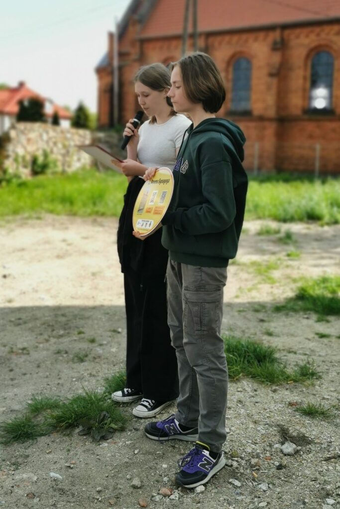 Fotografia przedstawia dwójkę uczniów: chłopiec w zielonej bluzie trzyma w ręku żółtą, owalną tablicę na której widoczne są kody QR a obok niego stoi dziewczyna z mikrofonem w ręku. W tle obu postaci widoczny jest fragment ceglanego kościoła oraz kamiennego muru.