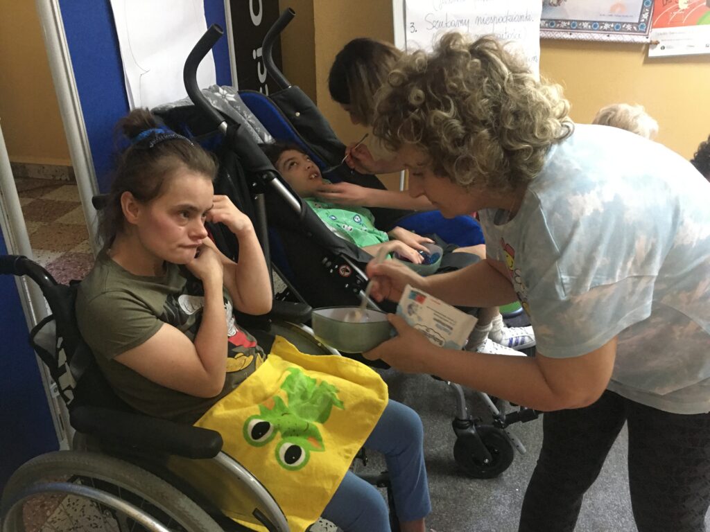 Korytarz szkolny. Na wózku inwalidzkim dziewczynka, a przed nią kobieta dająca jej z łyżeczki jedzenie.