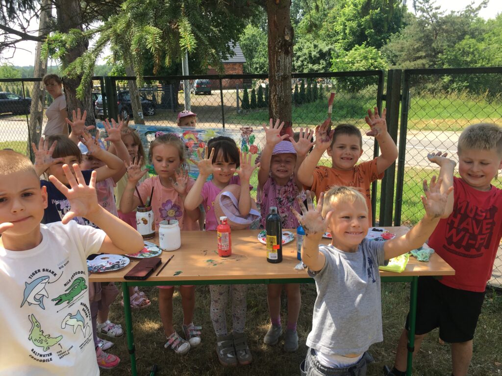 Grupa dzieci na dworze przed kolorowym obrazkiem. Wszyscy wznoszą do góry swoje kolorowe dłonie
