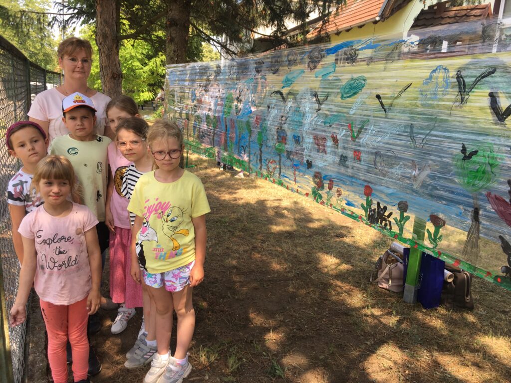 Podwórko szkolne, po lewej stronie stoi grupa dzieci, po ich prawej stronie pomalowana na kolorowo folia rozciągnięta między drzewami