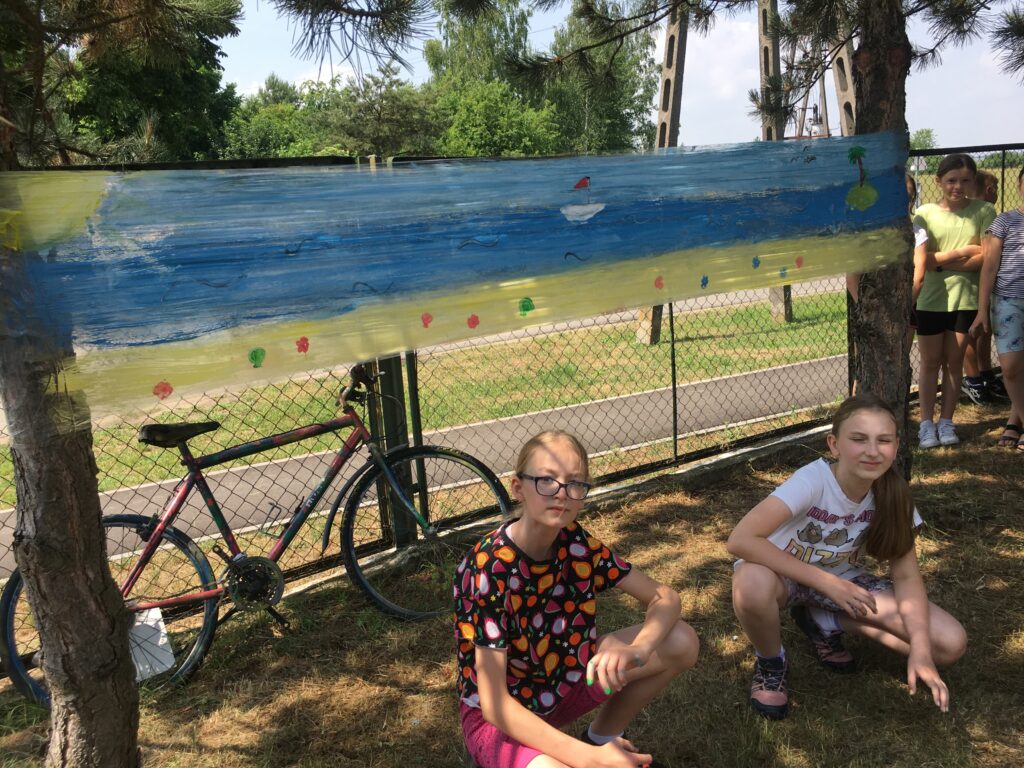 Podwórko szkolne, dwie dzieci kucają, na nimi pomalowana na kolorowo folia rozciągnięta między drzewami. W tle stoi rower.