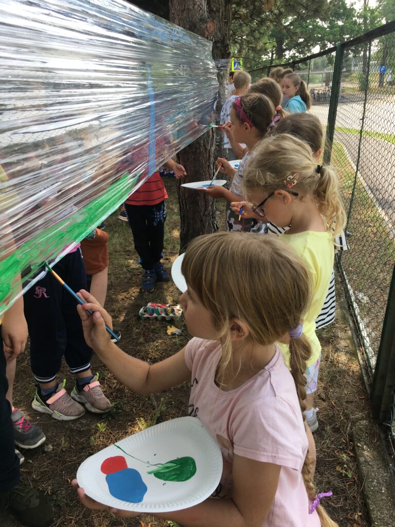 Podwórko szkolne, dzieci stoją pochylone,  w szeregu, przodem do powierzchni folii, rozciągniętej między drzewami, po której malują pędzlami. Stoją na tle pomalowanej na różnobarwne wzorki folii.
