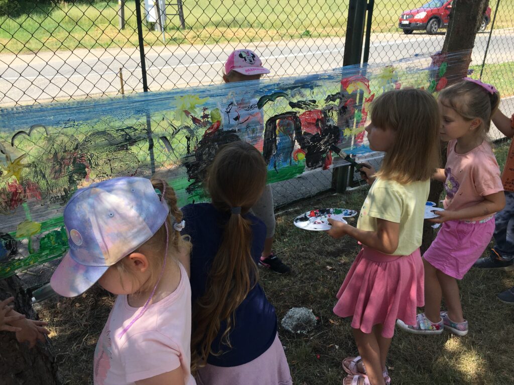 Podwórko szkolne, pięć dziewczynek stoi w szeregu przodem do powierzchni folii, rozciągniętej między drzewami, po której malują pędzlami. Stoją na tle pomalowanej na różnobarwne wzorki folii