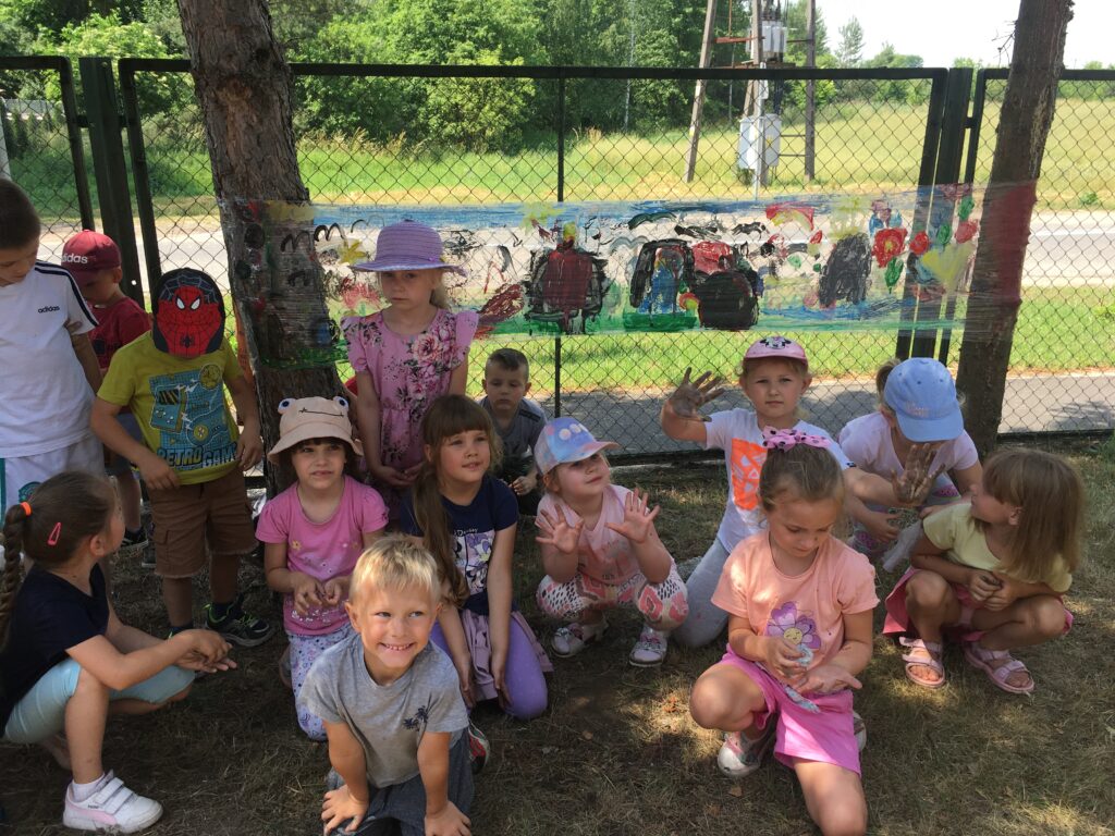 Grupa dzieci na dworze przed kolorowym obrazkiem. Wszyscy wznoszą do góry swoje kolorowe dłonie.