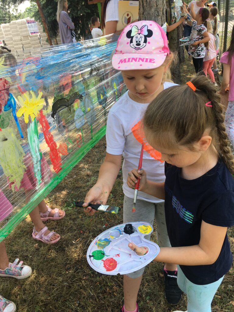 Podwórko szkolne, dwie dziewczynki stoją  przodem do powierzchni folii, rozciągniętej między drzewami, po której malują pędzlami. Trzymają w ręku paletę i pędzel. Stoją na tle pomalowanej na różnobarwne wzorki folii
