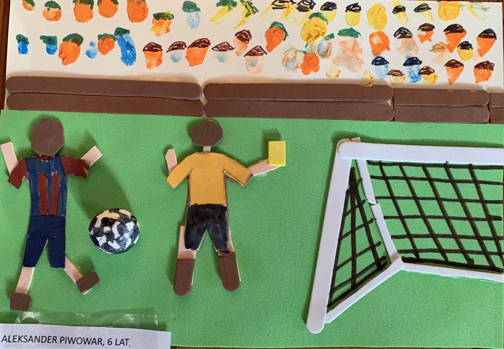 Praca 6 letniego chłopca przedstawia boisko, na którym piłkarz kopie piłkę a sędzia wystawia żółtą kartkę. Obok wyklejona jest bramka, praca wyklejona jest z miękkiego materiału. W górnej części pracy znajdują się trybuny, głowy ludzi wykonane są z odciśniętych palców.