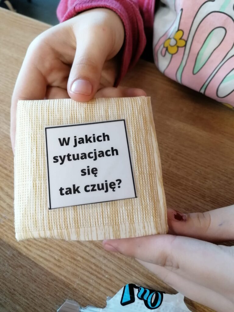 Na środku kwadratowa poduszeczka z kwadratowym białym obrazkiem z czarnym kolorem zapisanym zdaniem –„w jakich sytuacjach się tak czuję-”. Poduszeczkę trzymają dłonie dziecka