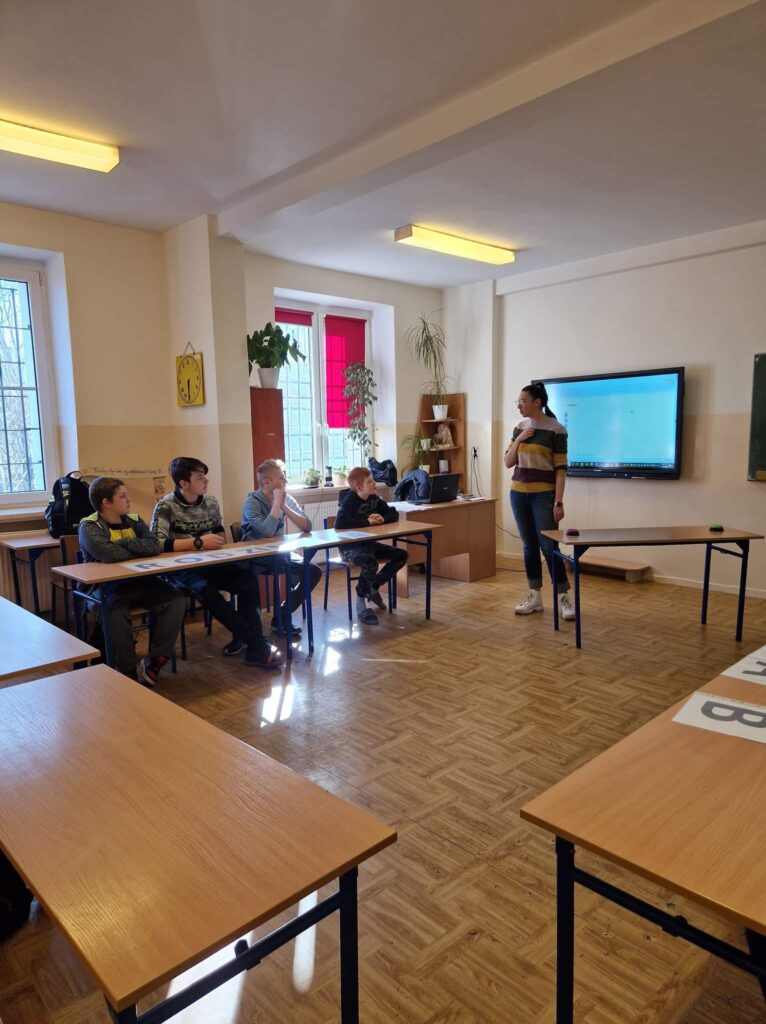 Na zdjęciu widoczny doradca - nauczyciel prowadzący lekcję, w ławkach siedzą uczniowie. Słuchają co nauczyciel mówi. 
