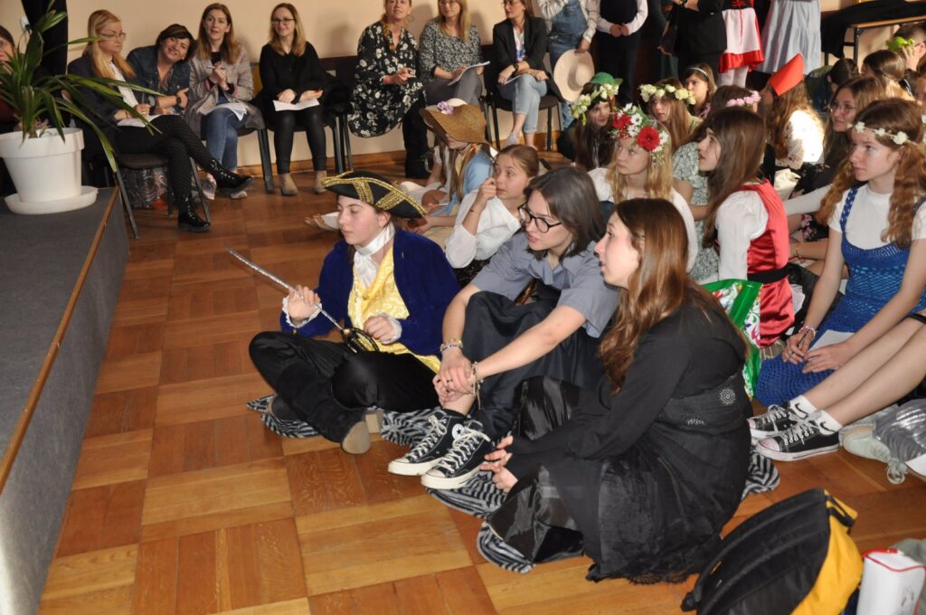 Sala, na środku obok siebie siedzą na podłodze dzieci w kolorowych strojach