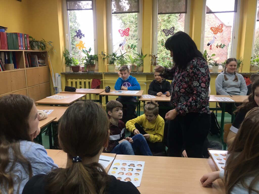 Sala lekcyjna, stoliki ustawione w kształcie kwadratu, siedzą w nich dzieci.. Na środku na poduszkach , na podłodze  siedzą dzieci. Ponad nimi stoi nauczycielka i coś pokazuje. Na pulpicie leżą plansze z emocjami – twarzami. W tle okna z naklejonymi kolorowymi motylami.