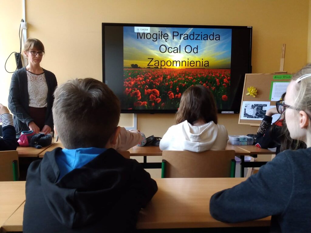 Sala lekcyjna, uczniowie siedzą w ławkach i 
Patrzą na wyświetlany na tablicy obrazek. Przy ekranie stoi nauczyciel – kobieta w czarnym sweterku.
