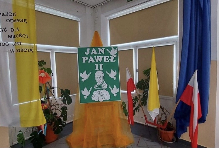Wystawa poświęcona Papieżowi. Na środku wizerunek Jana Pawła II na cokole. Po prawej stronie biało – czerwone flagi oraz żółta flaga. Po lewej stronie na żółto- białym tle napis.