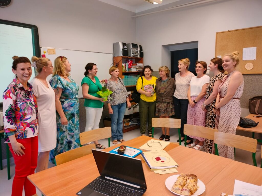 Sala lekcyjna. Na wprost stoją kobiety, w tyle meble. Jedna z kobiet trzyma kwiat a inna torebkę. Na środku stół a na nim rozłożony laptop.

