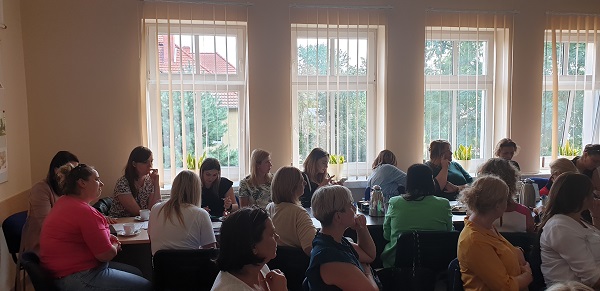 Sala konferencyjna, przy stołach siedzą uczestnicy szkolenia. Za  nimi okna z pionowymi roletami. 

