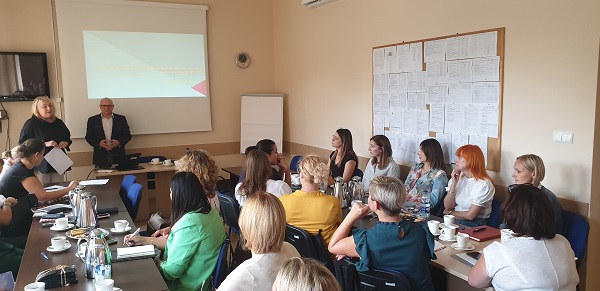 Sala konferencyjna, na środku stoły ustawione w kształcie prostokąta. Wokół siedzą nauczyciele. Przed nimi stoją dwie osoby. W tle ekran z wyświetloną prezentacją.

