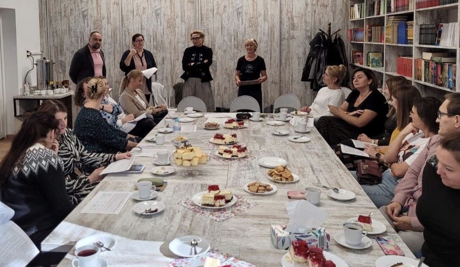 Sala biblioteczna, na środku stoły ustawione w kształcie prostokąta. Wokół siedzą nauczyciele. Przed nimi stoją trzy kobiety i jeden mężczyzna. Po prawej stronie regały z książkami. Na stole stoją talerzyki z ciastem i białe filiżanki. 