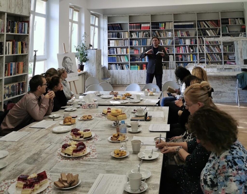 Sala biblioteczna, na środku stoły ustawione w kształcie prostokąta. Wokół siedzą nauczyciele. Za nimi stoi w rozkroku na tle regałów z książkami jeden mężczyzna i trzyma w ręku książkę. Po prawej stronie regały z książkami. Na stole stoją talerzyki z ciastem i białe filiżanki. 