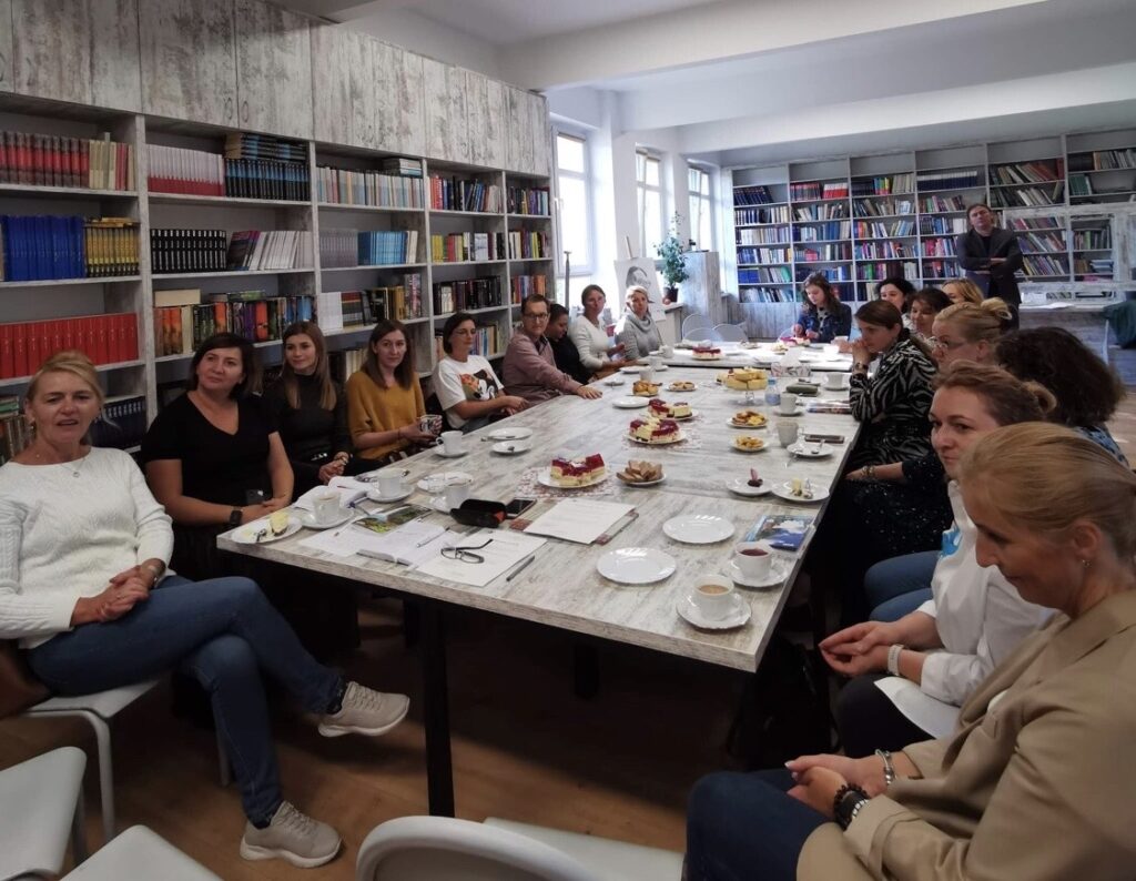 Sala biblioteczna, na środku stoły ustawione w kształcie prostokąta. Wokół siedzą nauczyciele. Za nimi stoi na tle regałów z książkami mężczyzna. Po prawej stronie regały z książkami. Na stole stoją talerzyki z ciastem i białe filiżanki. 

